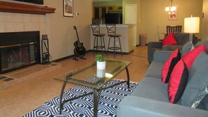 Un salón con una mesa de centro de vidrio, un sofá con cojines rojos, una chimenea, una guitarra y una zona de cocina al fondo.