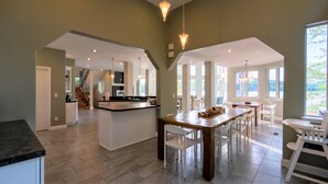 Una cocina amplia con una isla grande, un comedor con sillas blancas y vistas al exterior a través de ventanas grandes.
