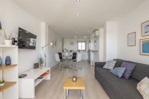 A modern living room with a sofa, coffee table, and TV. There is a dining area with chairs and a table, and a kitchen in the background.