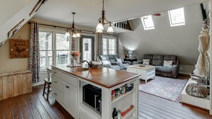 Una cocina acogedora con una isla central, armarios blancos y un techo rústico. Hay una zona de comedor con un sofá de cuero y una pequeña mesa.