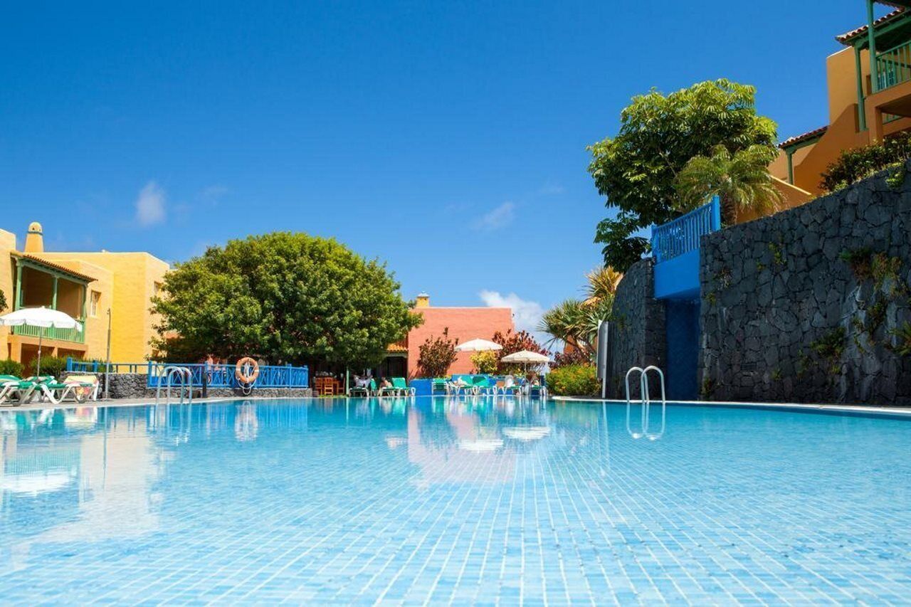 Ein klares, blaues Schwimmbecken mit Liegestühlen und Sonnenschirmen, umgeben von Gebäuden und einem großen Baum.