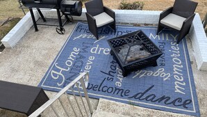 Una terrazza esterna con una griglia per barbecue, due poltrone in vimini e un braciere su un tappetino blu con scritte.