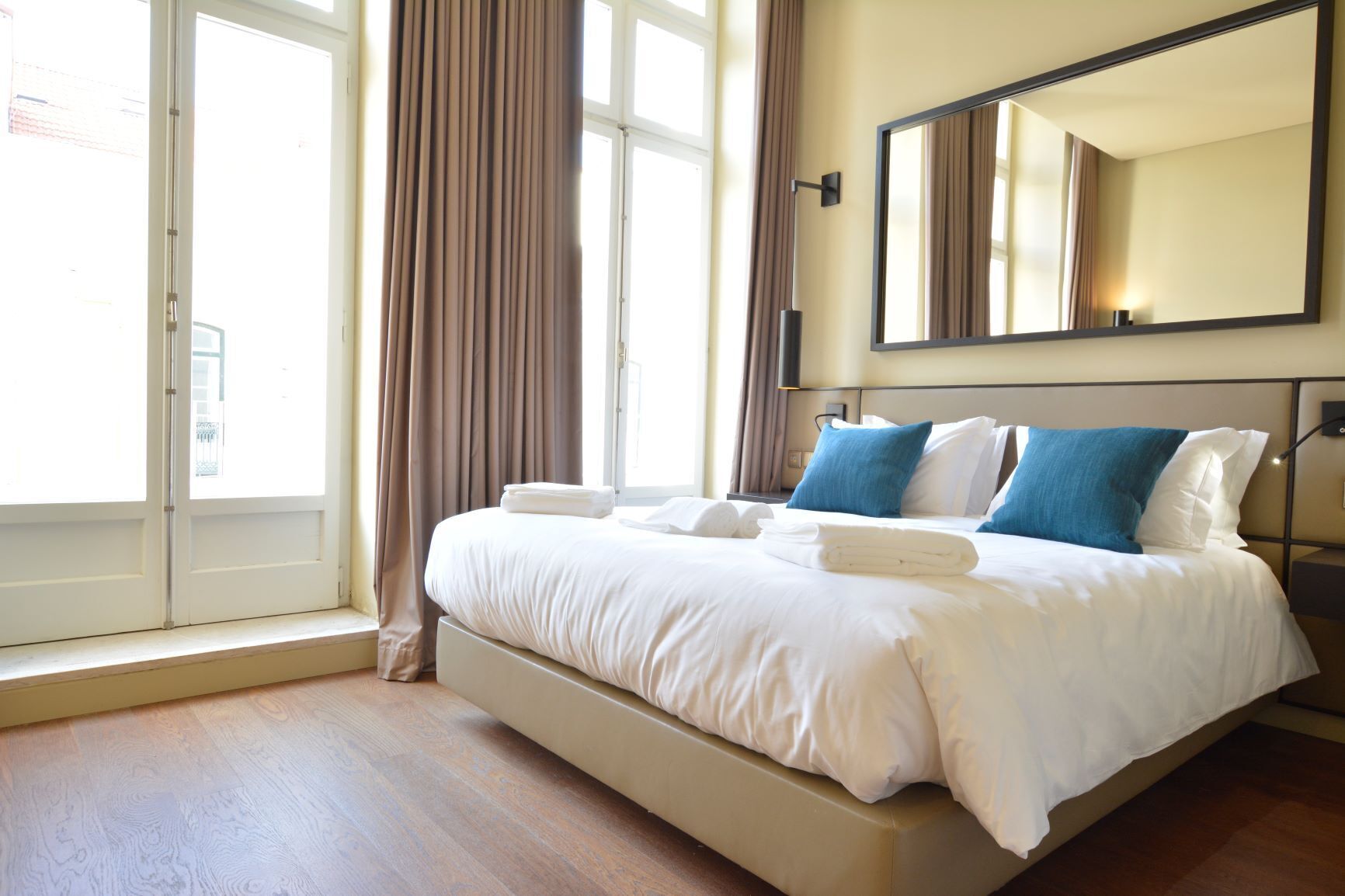 A hotel room with a large bed, a window with curtains, a wall-mounted mirror, and a bedside table with a lamp.