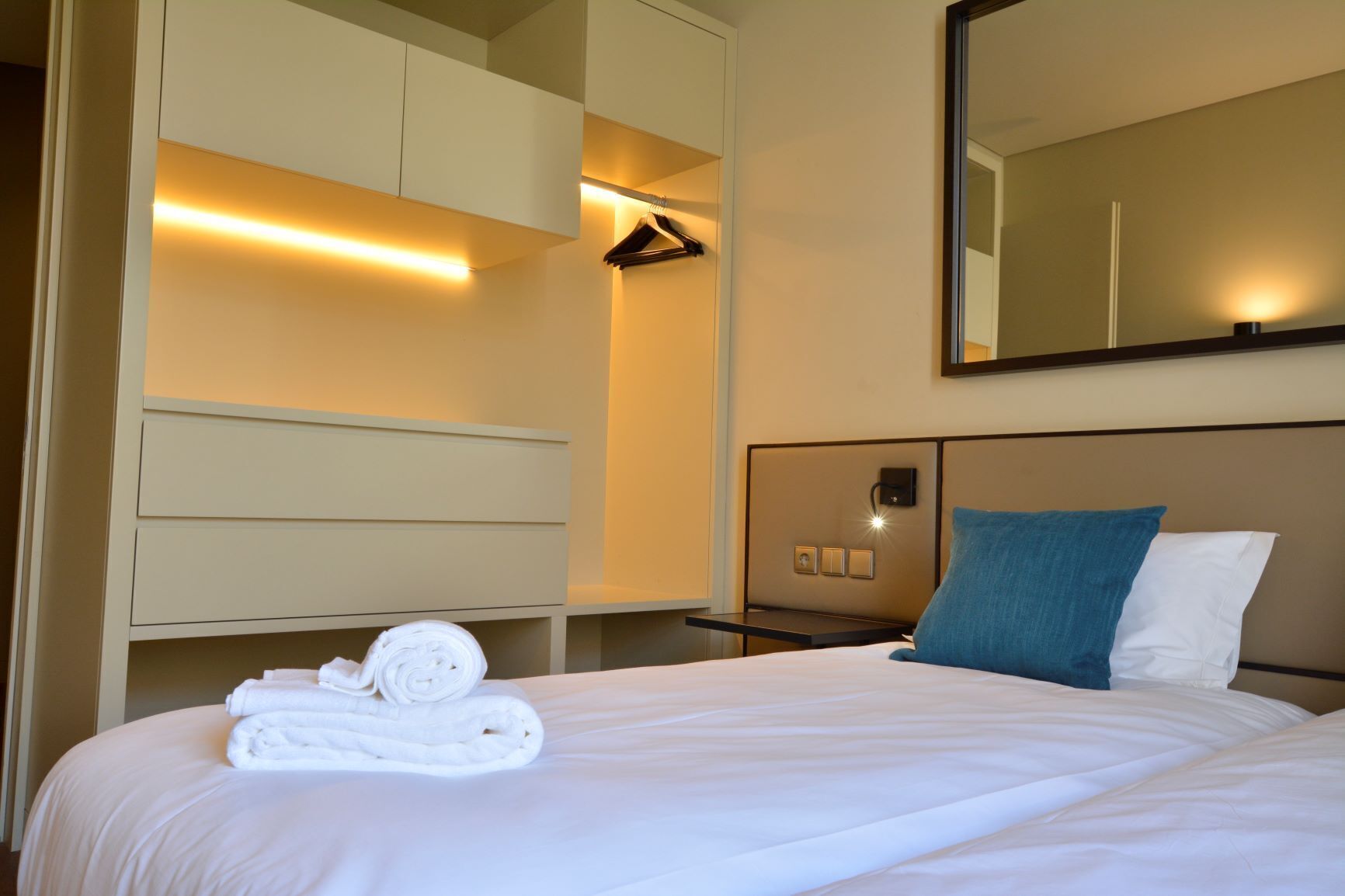 A hotel room with a neatly made bed, a hanging closet, a bedside table with a lamp, and a large mirror on the wall.