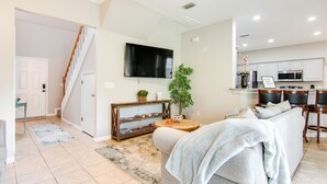 A modern living room with a sofa, a coffee table, and a TV mounted on the wall. There is a staircase leading to another room, and a kitchen area with cabinets and appliances. - Serene - Fully Furnished Immaculate with 2-car Garage! (Lutz)