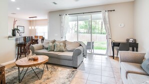 A spacious living room with a grey sofa, a coffee table, and a dining area with chairs and a table. - Serene - Fully Furnished Immaculate with 2-car Garage! (Lutz)
