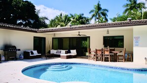 Una piscina exterior con borde de azulejos, un área de estar techada con sofá y sillones, y una parrilla.