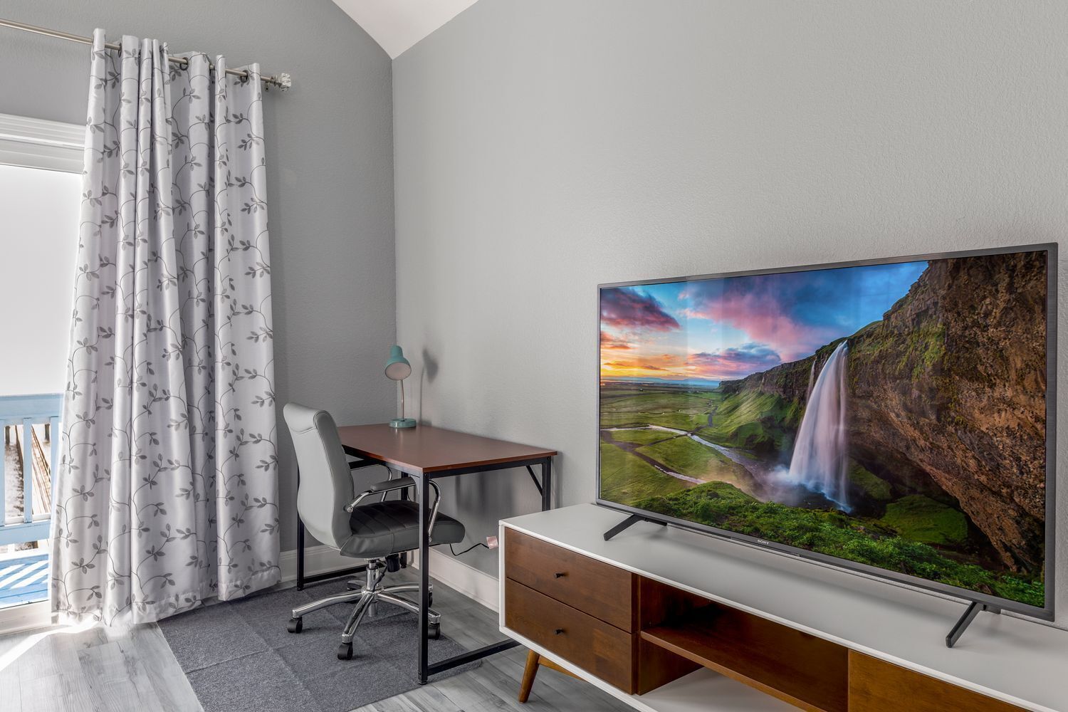 A hotel room with a desk, chair, TV, and a view of a waterfall.