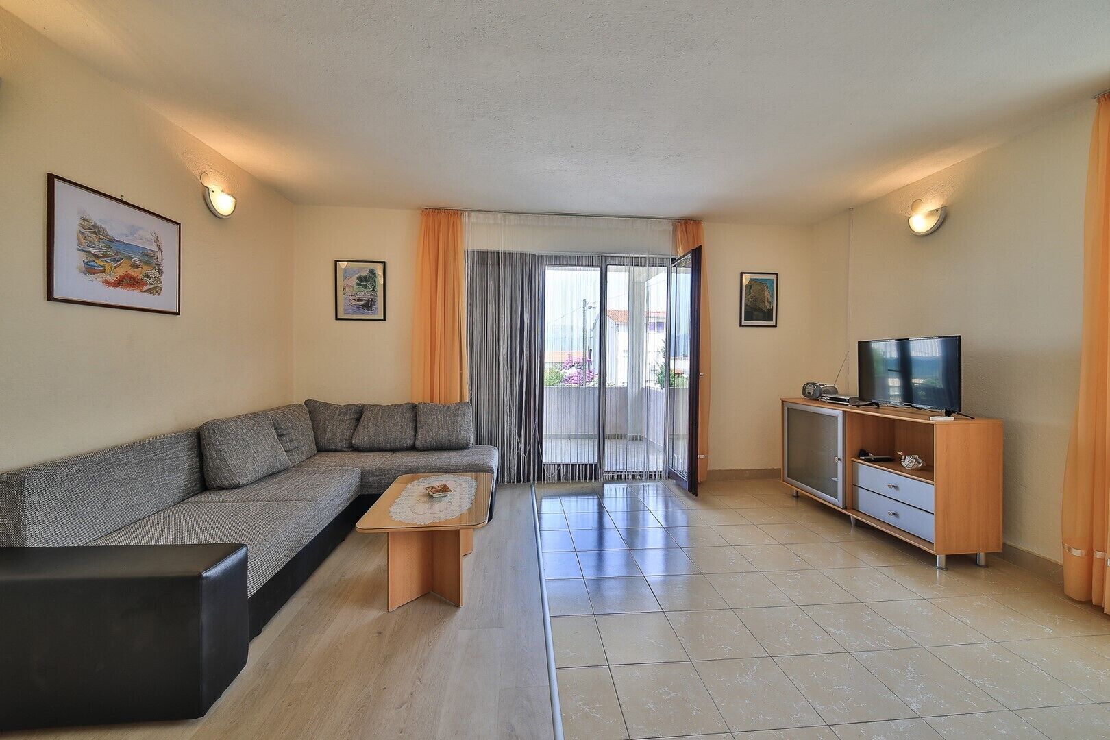 Una sala de estar con un sofá, una mesa de centro, un televisor sobre un mueble de madera y un balcón con cortinas.