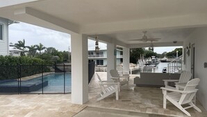 Un espacio exterior techado con piscina, sillas blancas y vistas a un puerto deportivo.