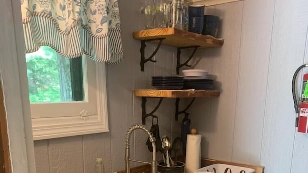 Ett kompakt kök med en vit diskho, en bänkskiva i trä och vita skåp. Ovanför diskhon finns hyllor där tallrikar och glas förvaras.