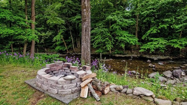 A stone fire pit surrounded by logs and rocks, set in a grassy area with a stream and dense forest in the background. - Cabin by the Creek with private creek access - (Tannersville)