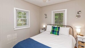 Un dormitorio con una cama, dos mesitas de noche, un ventilador de techo y dos ventanas con persianas.