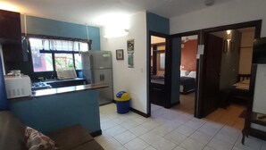 Een kleine keuken met een koelkast, magnetron en wastafel. Er staat een kleine prullenbak en een raam met zonwering. De vloer is betegeld en er is een deur naar een slaapkamer met een bed en nachtkastje.