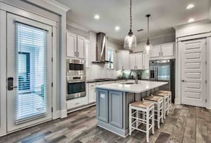 A modern kitchen with white cabinetry, stainless steel appliances, and a central island with bar stools. - Sea-Struck, Elegant 3BR/3BA w/6 seater golf cart! (Santa Rosa Beach)