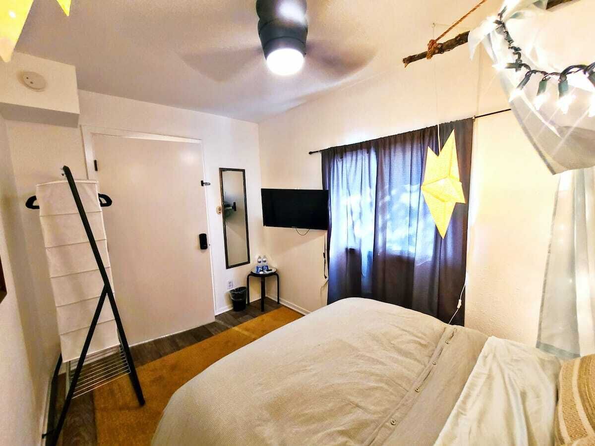 A hotel room with a bed, a ceiling fan, a flat-screen TV, a small table with a chair, and a window with curtains.