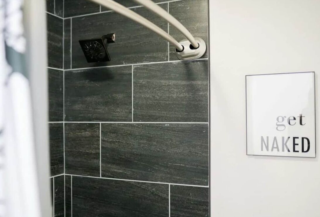 A bathroom with dark tiles and a shower head. There is a sign on the wall that says "get NAKED".