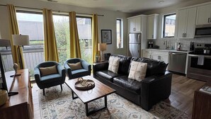 Een moderne woonkamer met een bank, fauteuils, een salontafel en een keukenhoek met kasten en apparatuur.