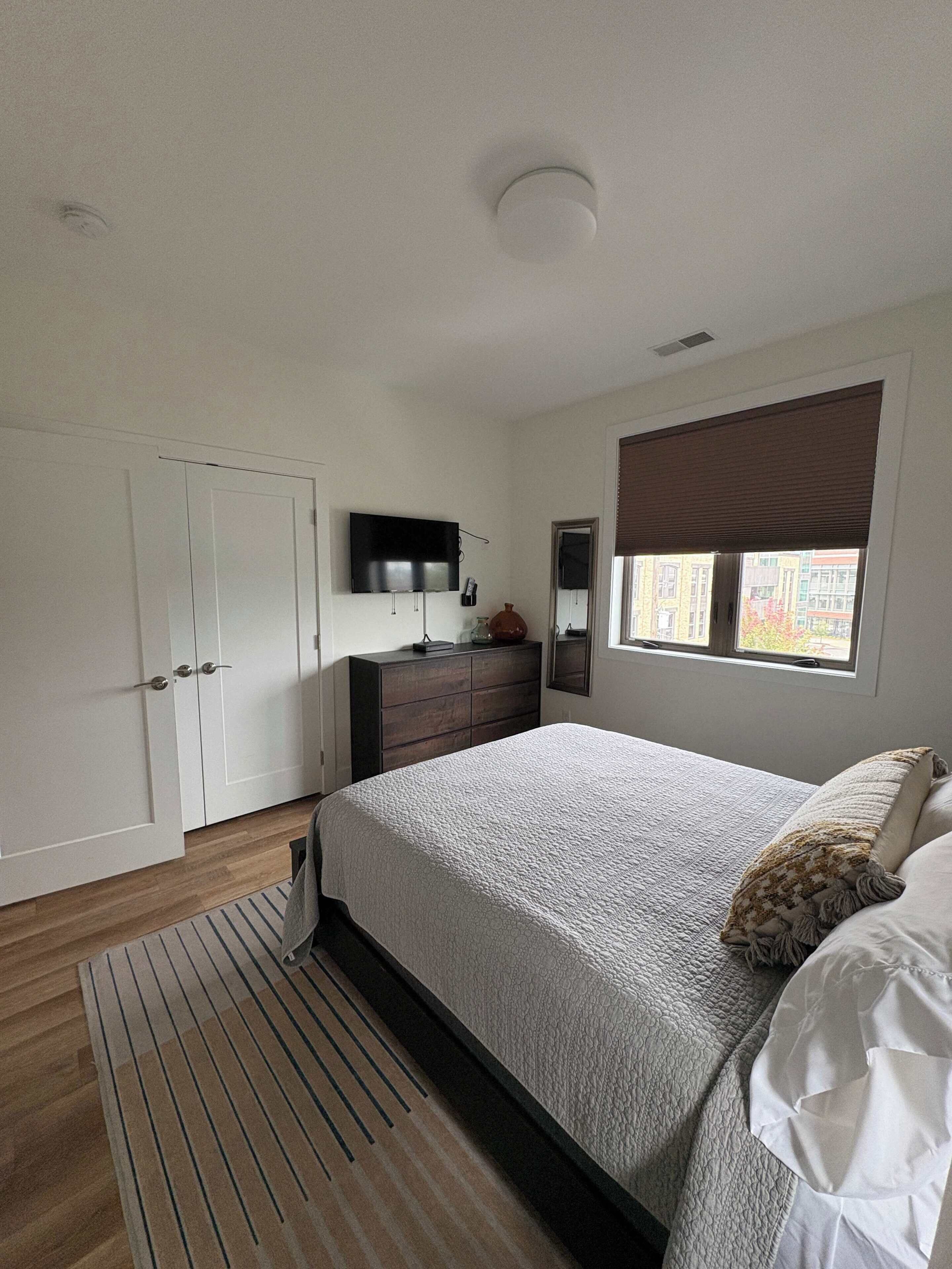 Une chambre à coucher comprenant un lit, une commode, un téléviseur, une fenêtre avec des stores et une porte.