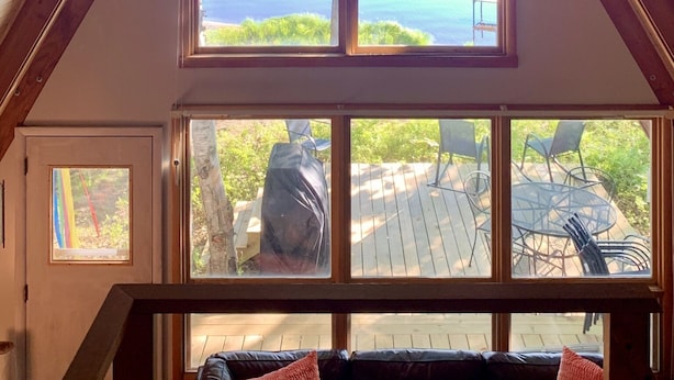 A room with a sloped ceiling, wooden beams, and large windows offering a view of the sea. - A-Frame cabin located on the shores on lake Independence. (Big bay)