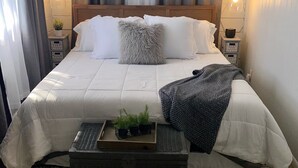 A neatly made bed with white linens, a gray fluffy pillow, and a black and white patterned throw. There is a small wooden side table with a plant and a lamp. The room has a patterned rug and a window with curtains. - Top-Rated Balsam Lake Vacation Home with Stunning Lake Views, 4 BR & Family Fun (Balsam Lake)