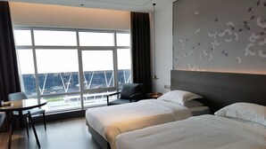 A hotel room with two beds, a desk, and a view of a cityscape through large windows. - Dazhong Airport Hotel (Shanghai)