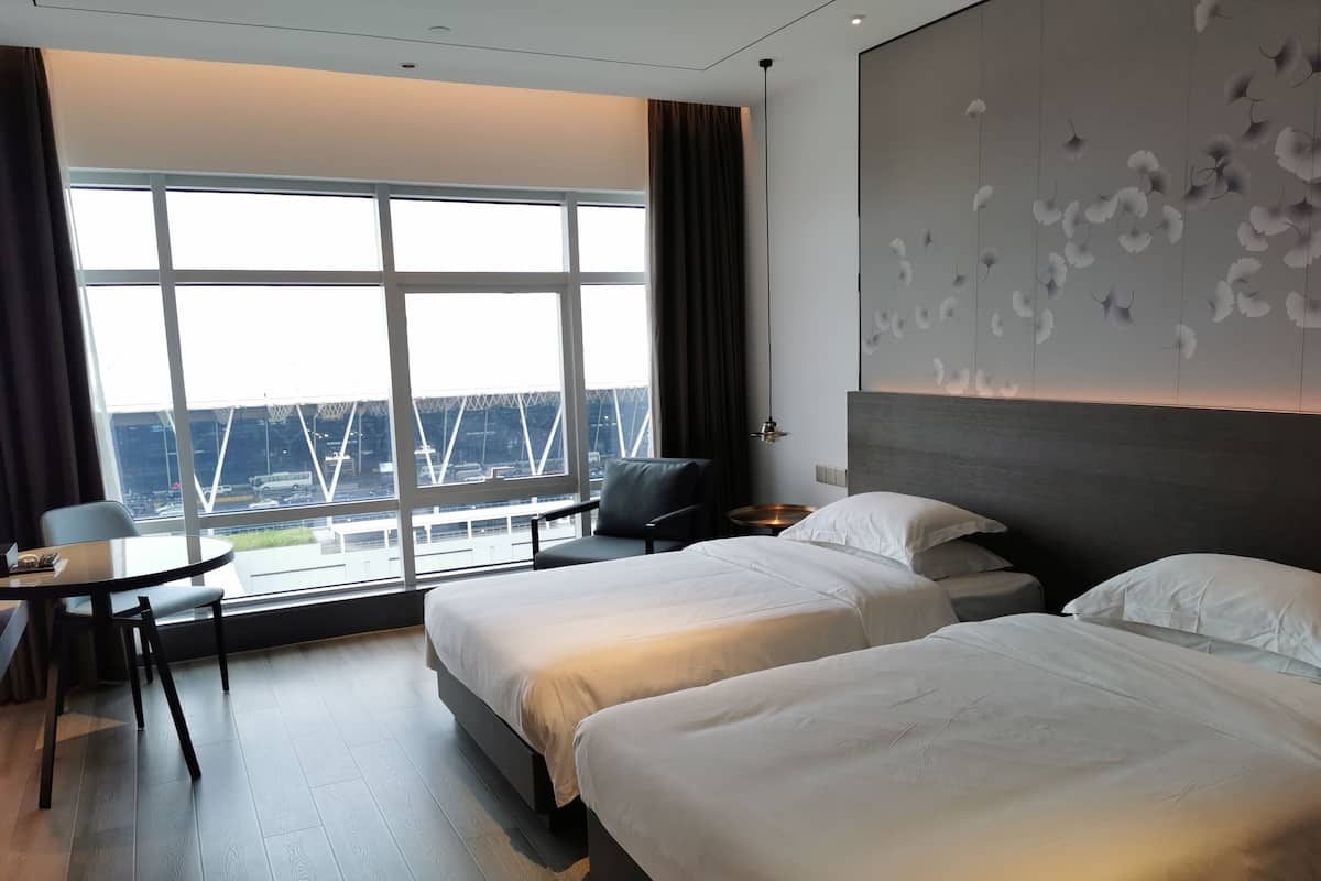 A hotel room with two beds, a desk, and a view of a cityscape through large windows.