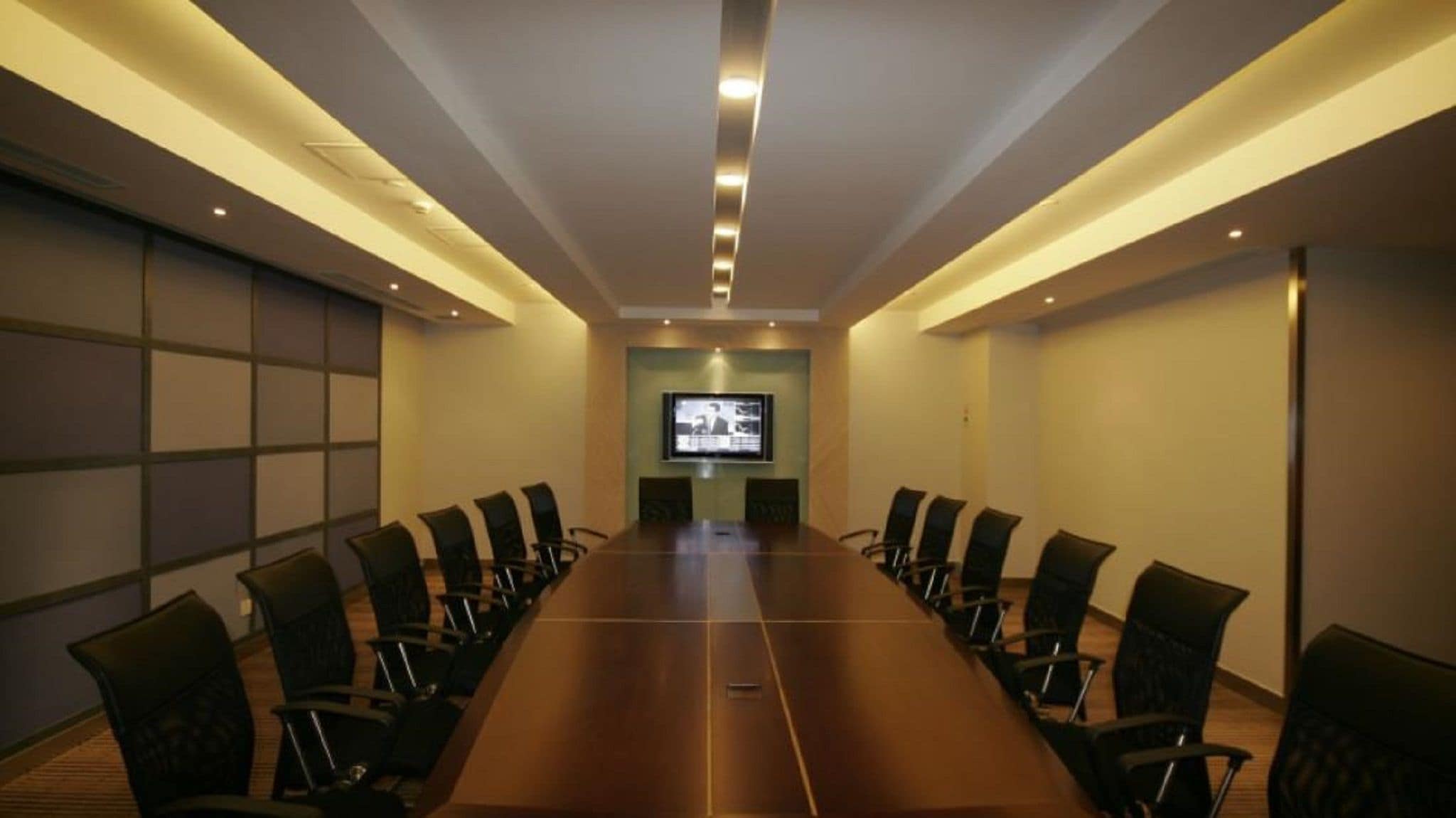 A conference room with a long table and chairs, a television screen, and recessed lighting.