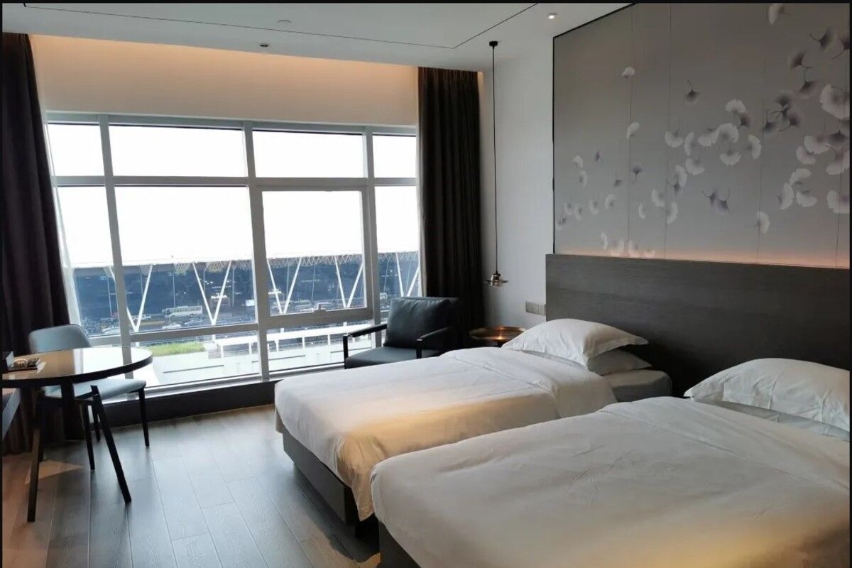 Habitación de hotel con dos camas, un escritorio y un ventanal grande con vistas a un puente.