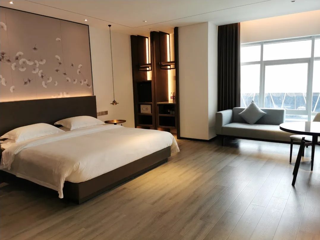 Une chambre d’hôtel moderne, dotée d’un grand lit, d’un canapé, d’une petite table et offrant une vue sur la ville grâce à de grandes fenêtres.