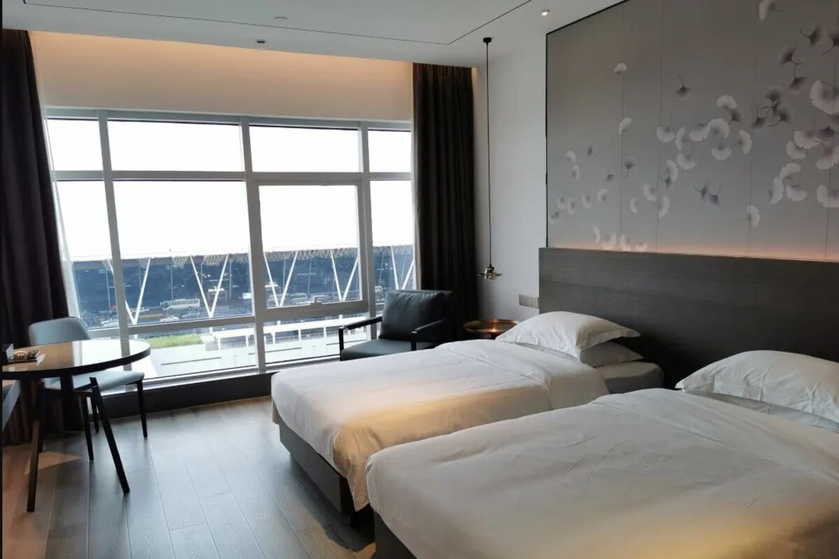 A hotel room with two beds, a desk, and a large window offering a view of a bridge.