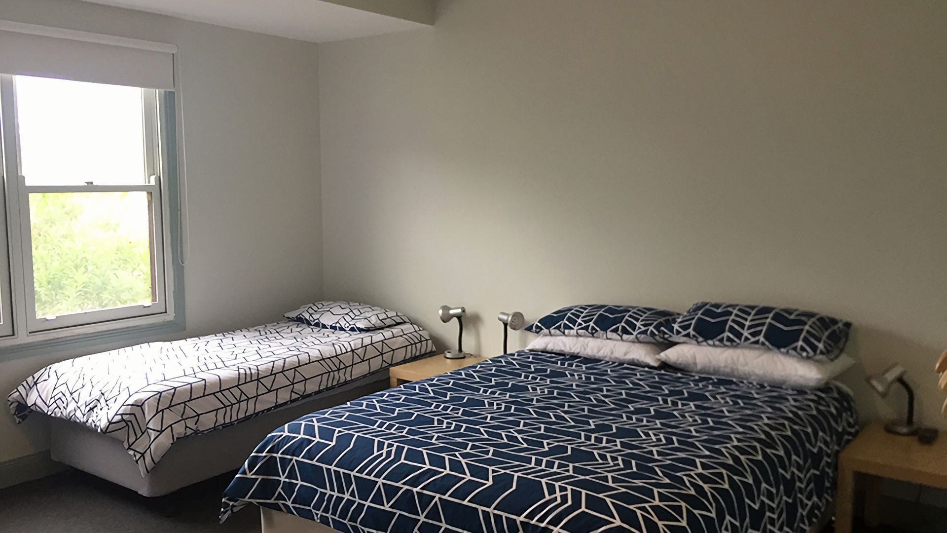 A bedroom with two beds, one with a geometric patterned bedspread and the other with a solid color. There is a window providing natural light, and a bedside table with a lamp.
