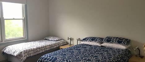 A bedroom with two beds, one with a geometric patterned bedspread and the other with a solid color. There is a window providing natural light, and a bedside table with a lamp.
