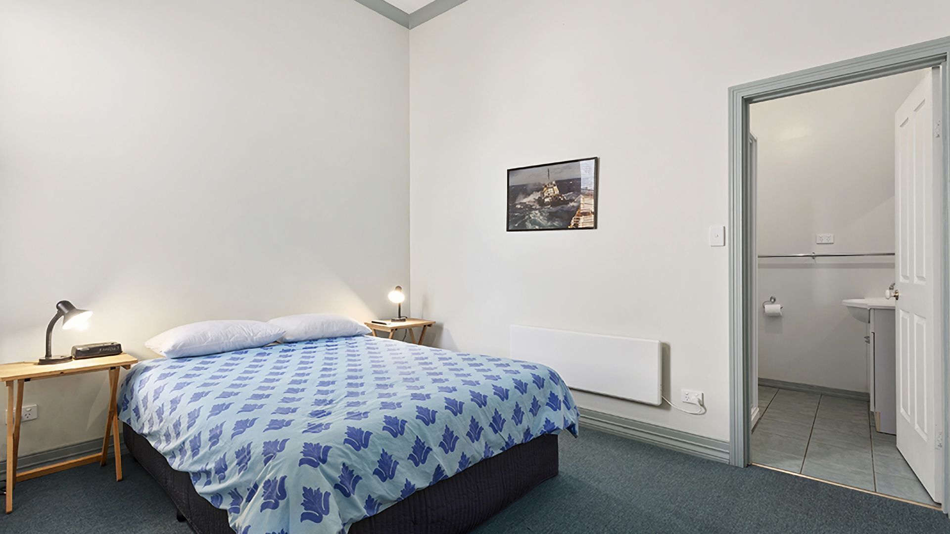 Une chambre à coucher avec un lit, deux tables de chevet avec des lampes, un radiateur, une porte donnant sur une salle de bain et un tableau encadré au mur.