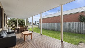 A covered patio with a wooden deck, a black sofa, a wooden table, and a dining set. - Fishermans Cottage - Queenscliff (QUEENSCLIFF)