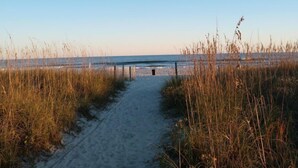 A sandy beach path leading to a wooden bridge over water, surrounded by tall grasses.