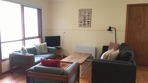 A living room with a sofa, a coffee table, and a TV. There is a window with curtains, a radiator, and a door. - The Beach Shack - Queenscliff (Queenscliff)