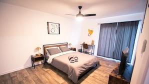 A bedroom with a bed, a desk, a chair, a ceiling fan, and a window with curtains. - Atomic Age (Austin)