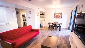 A living room with a red sofa, a wooden coffee table, a dining area with a table and chairs, and a kitchen in the background. - Atomic Age (Austin)