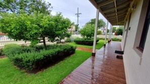 A covered porch with a brick floor, surrounded by well-maintained greenery and a white fence.