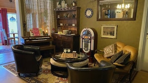 Uma sala de estar acolhedora com um piano, poltronas de couro, uma mesa de centro com um jogo de xadrez, uma estante de livros e um espelho na parede.