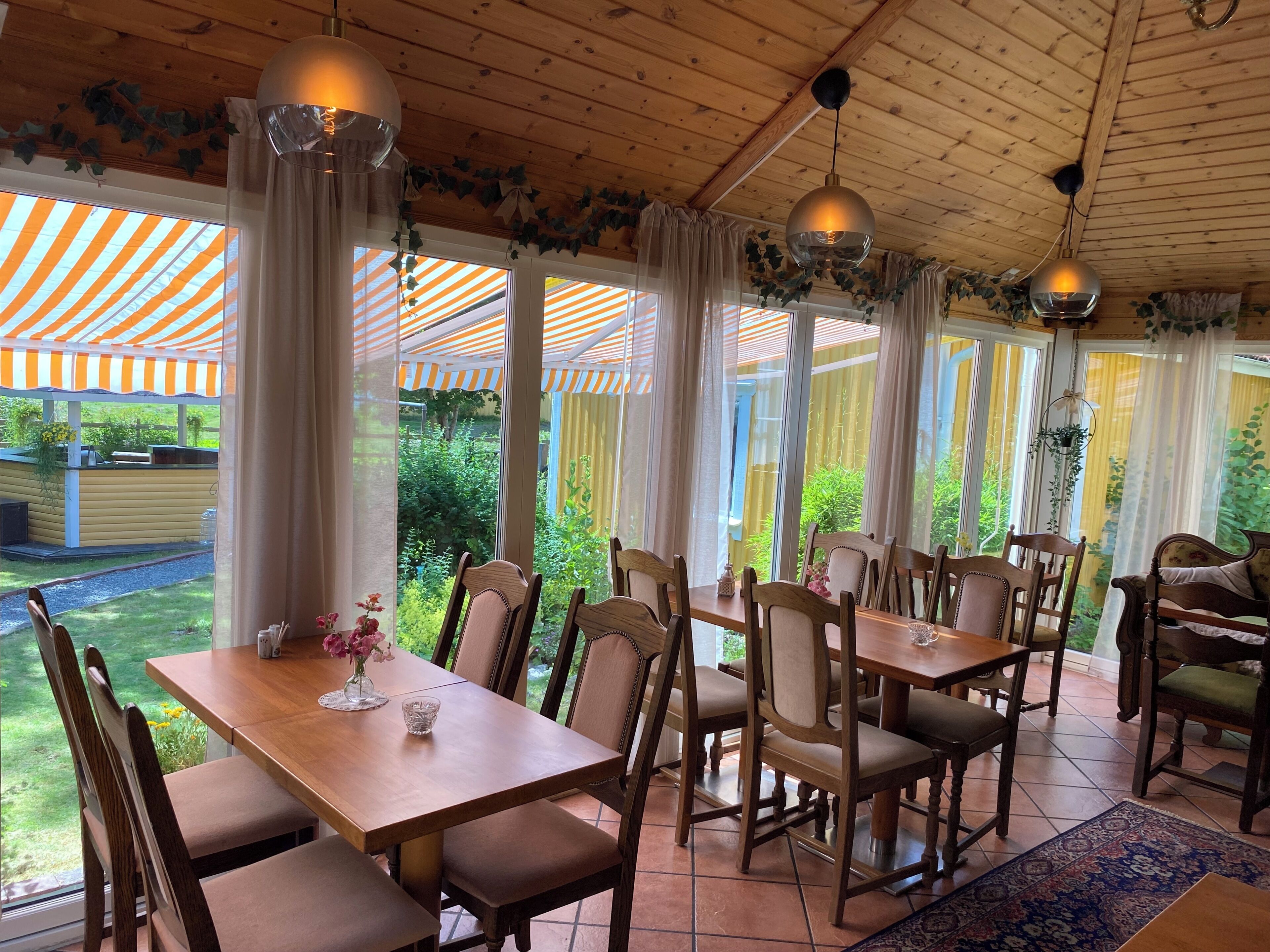 Een eethoek met houten tafels en stoelen, grote ramen met gordijnen en hangplanten.