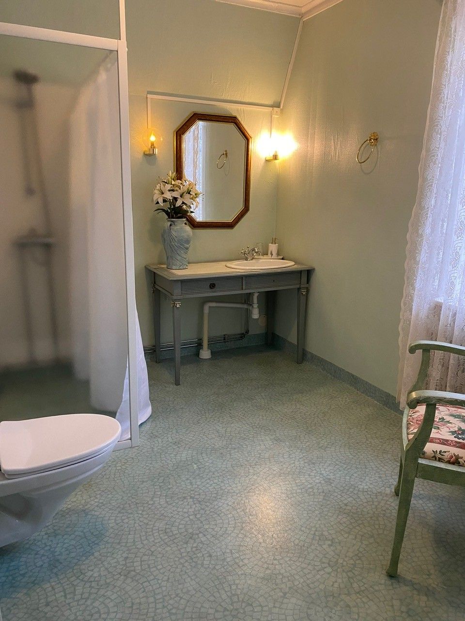 Un baño con ducha, inodoro y lavamanos. Hay un espejo sobre el lavamanos y un florero con flores en la encimera.