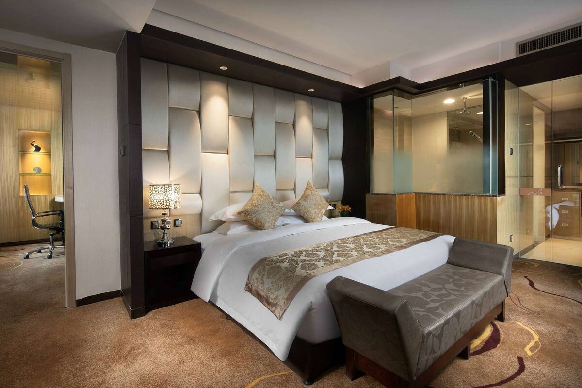A modern hotel room with a large bed, a sofa, a nightstand with a lamp, and a glass shower enclosure.