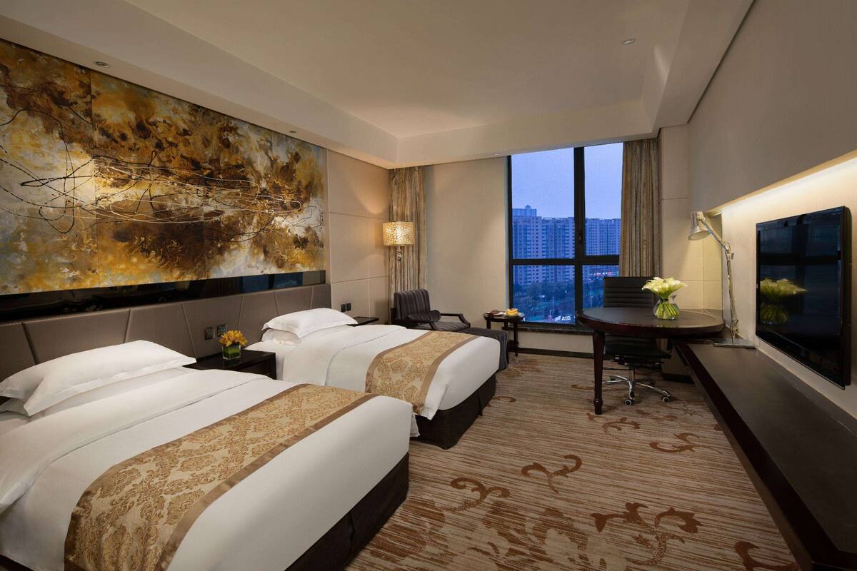 A hotel room with two beds, a large abstract painting, a desk with a lamp, and a view of buildings through the window.