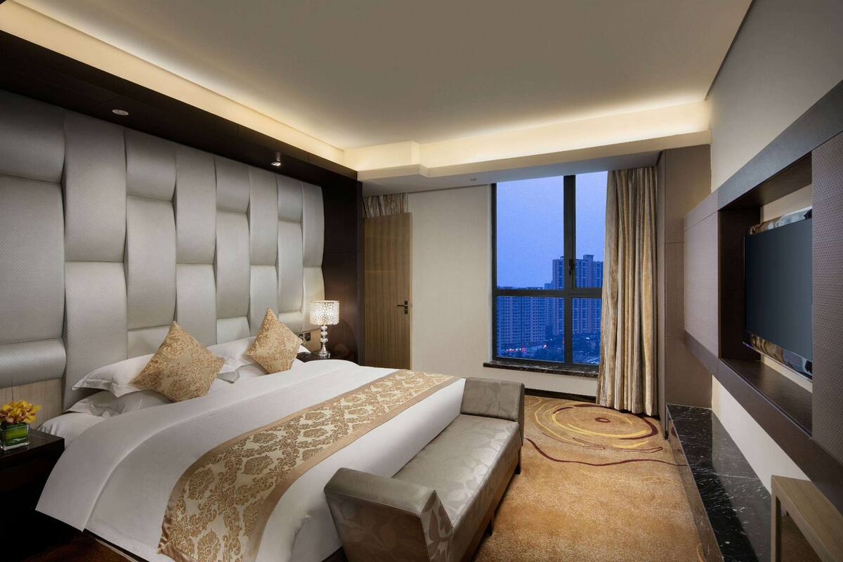 A modern hotel room with a large bed, a sofa, a TV, and a view of the cityscape.