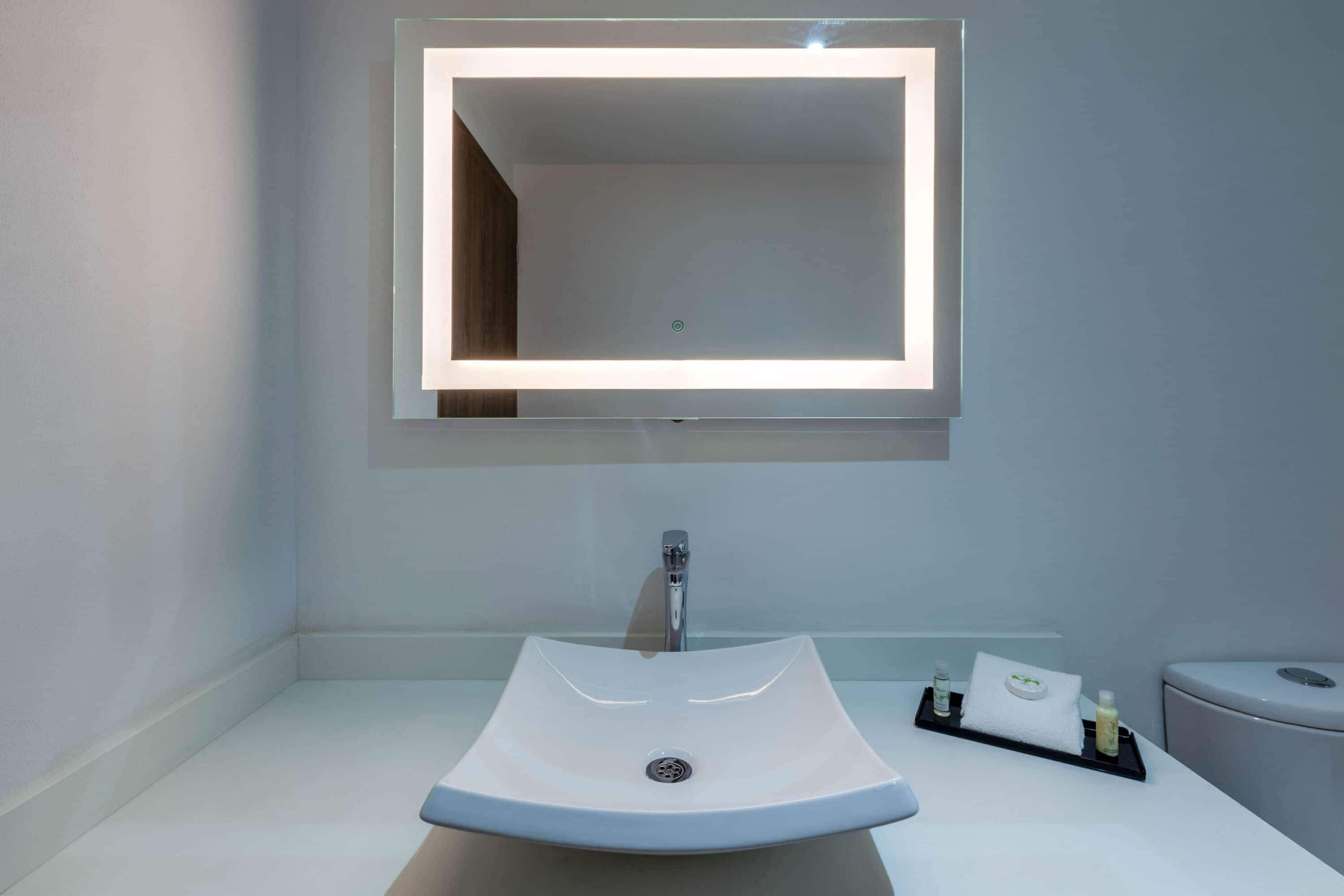 Un baño moderno con un lavamanos blanco, un espejo rectangular con iluminación integrada y una pequeña bandeja con artículos de aseo.