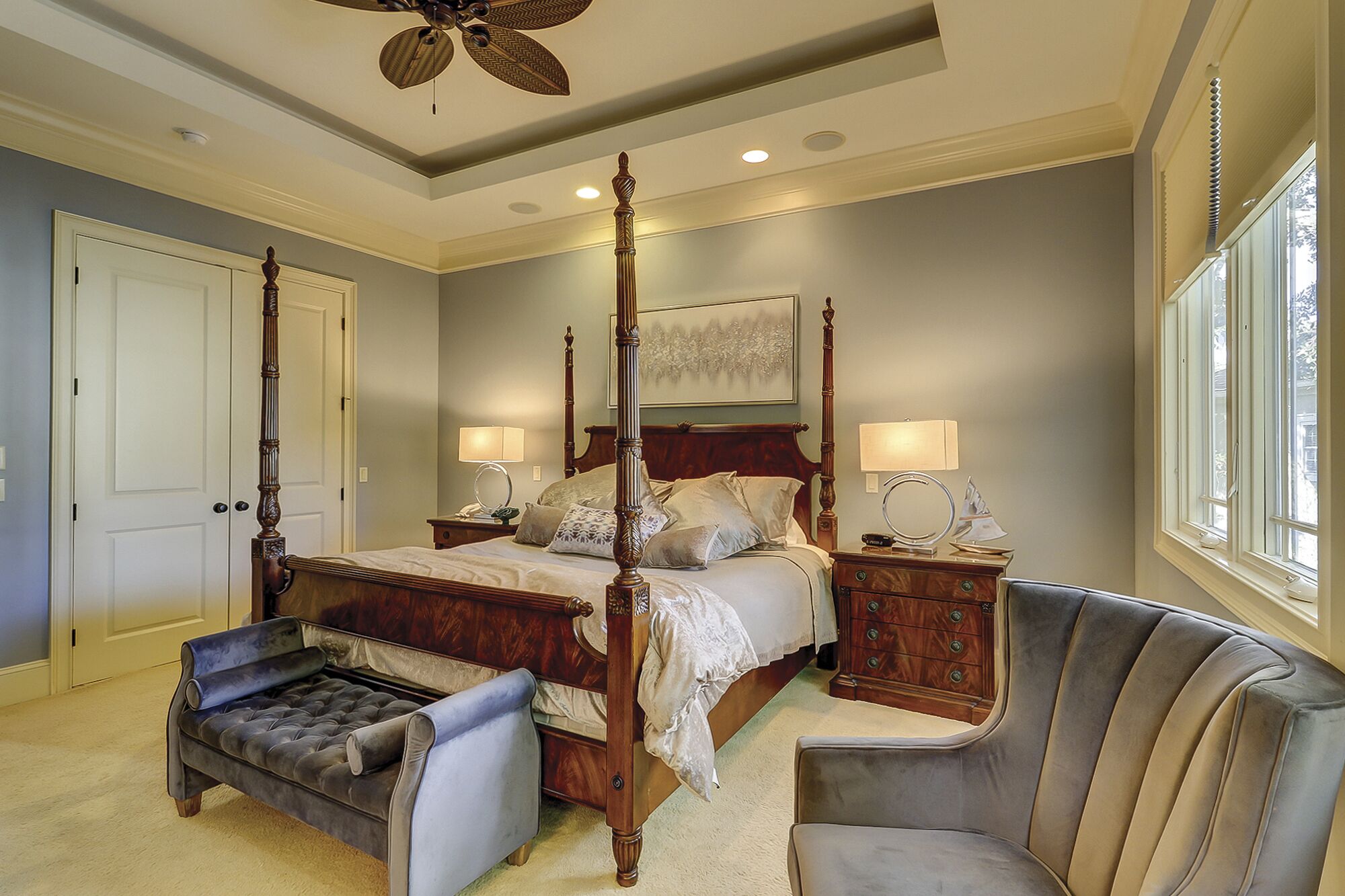 A four-poster bed with a wooden headboard, two bedside tables with lamps, a tufted armchair, and a large window with a view.