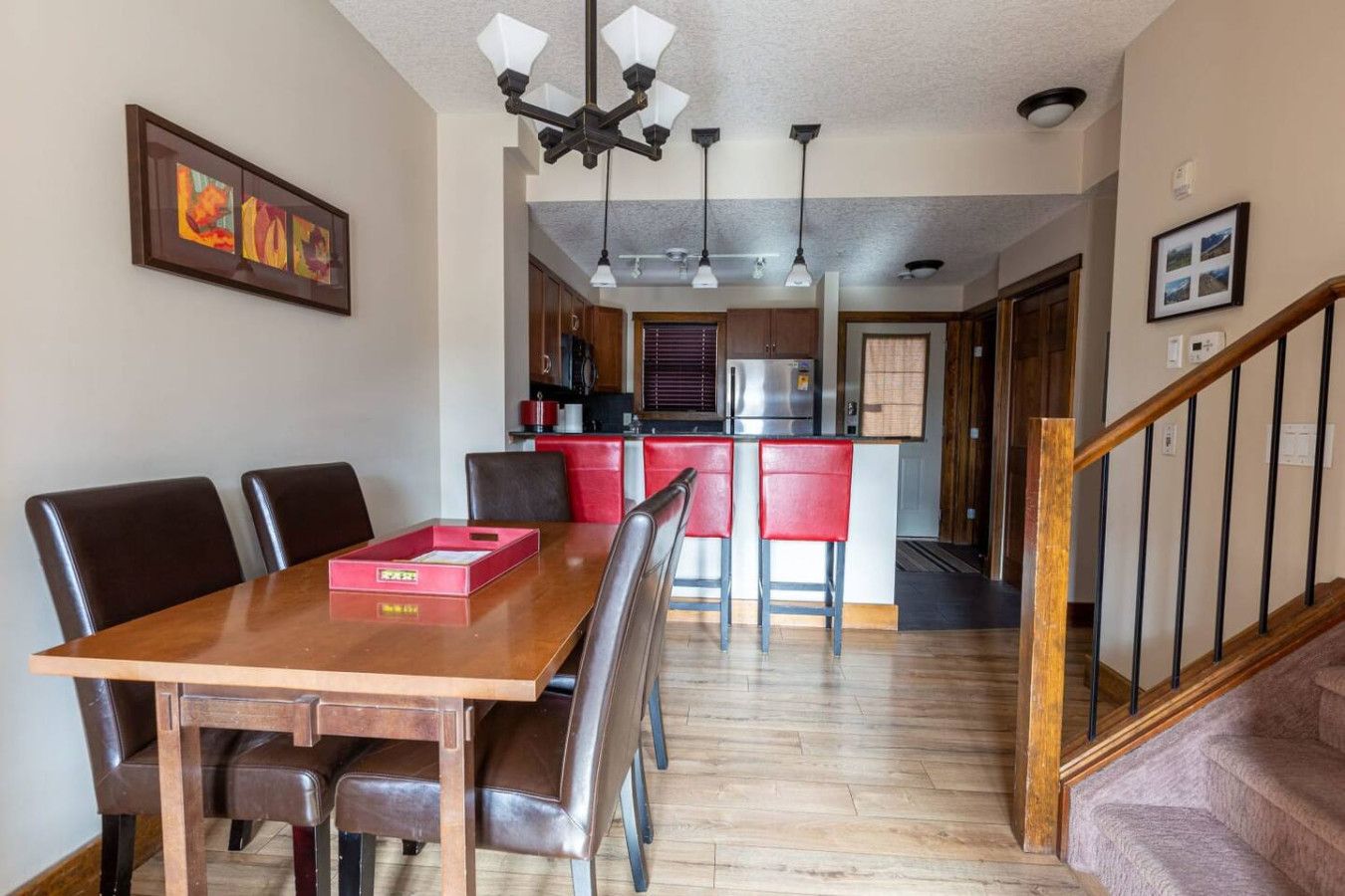 A dining area with a wooden table and chairs, a kitchen in the background, and a staircase leading to another level.