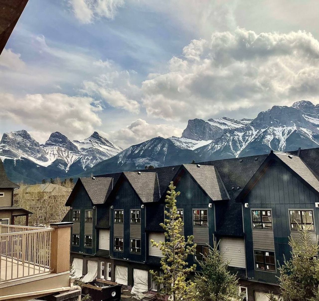 A row of dark wooden cabins with multiple windows, set against a backdrop of snow-capped mountains and a partly cloudy sky.